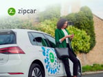 Woman in a green blazer using a smartphone next to a Zipcar vehicle parked in an urban setting, showcasing the convenience of car-sharing services.