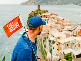 Man overlooking a picturesque coastal village while holding a flag that says "Get Your Guide," highlighting travel exploration and adventure.