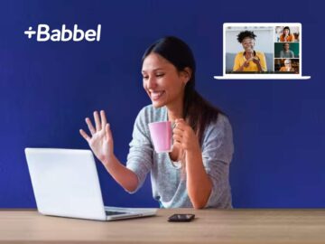 Woman engaging in an online language learning session with Babbel, smiling and holding a pink mug, while using a laptop on a wooden table with a blue background.