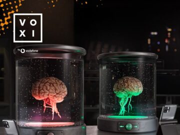 Image of two illuminated brain models in glass containers, one glowing red and the other green, set against a dark background with the VOXI logo in the top left corner. The display emphasizes innovative design and creativity in showcasing brain science or technology.