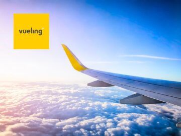 Aerial view from an airplane window showing clouds and blue sky with the Vueling Airlines logo in the corner.