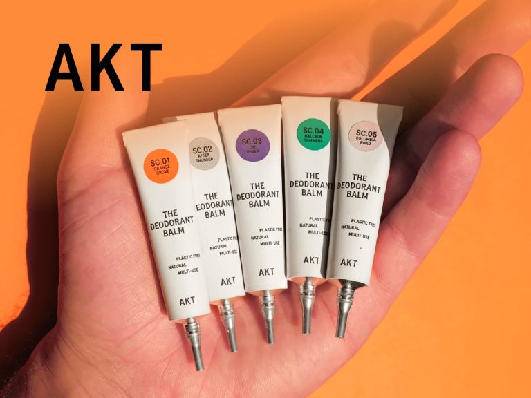 Hand Holding Four Akt Skincare Tubes Against An Orange Background, Showcasing A Variety Of Products For Skincare Routines.