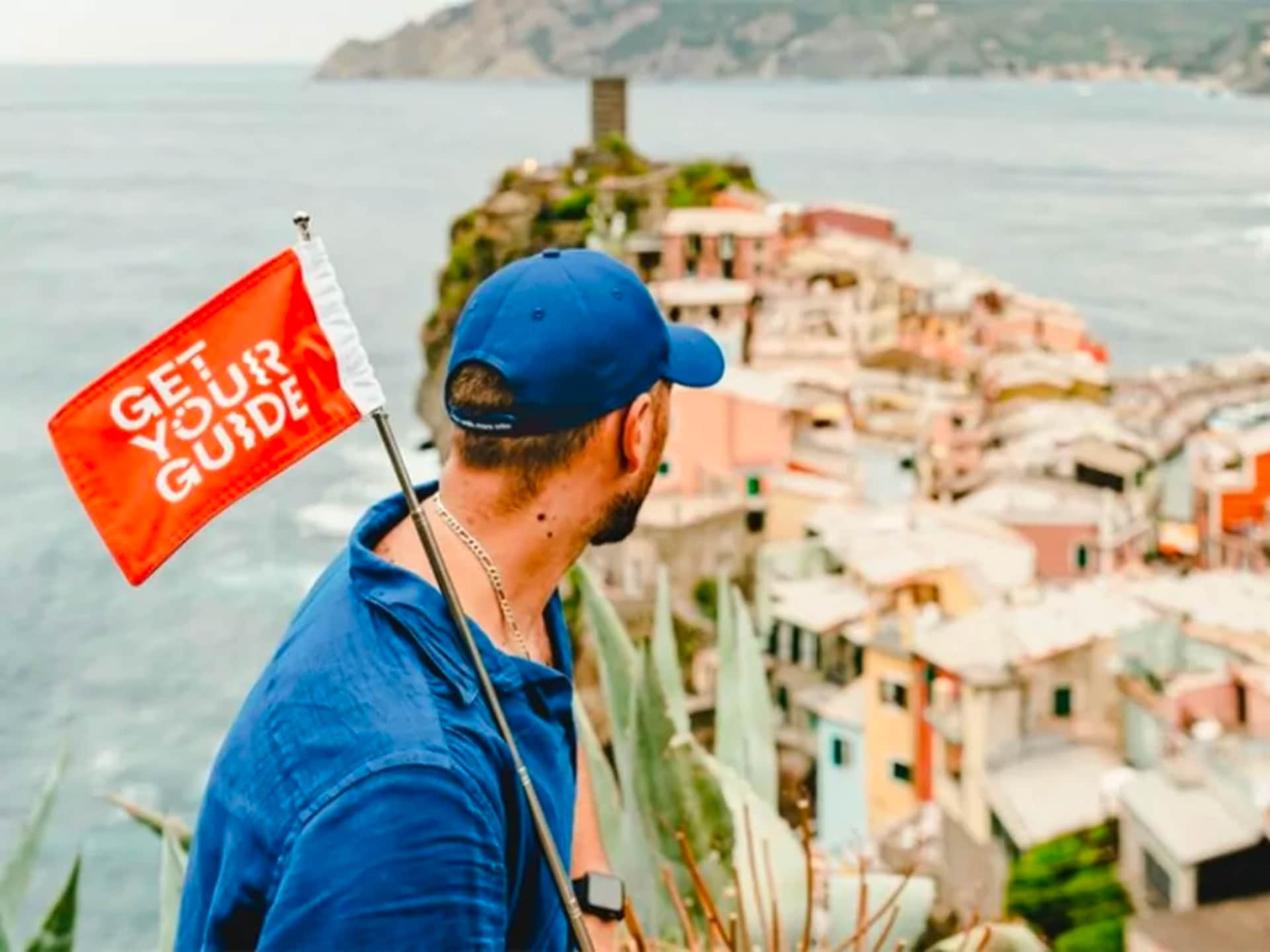 Man Overlooking A Picturesque Coastal Village While Holding A Flag That Says &Quot;Get Your Guide,&Quot; Highlighting Travel Exploration And Adventure.