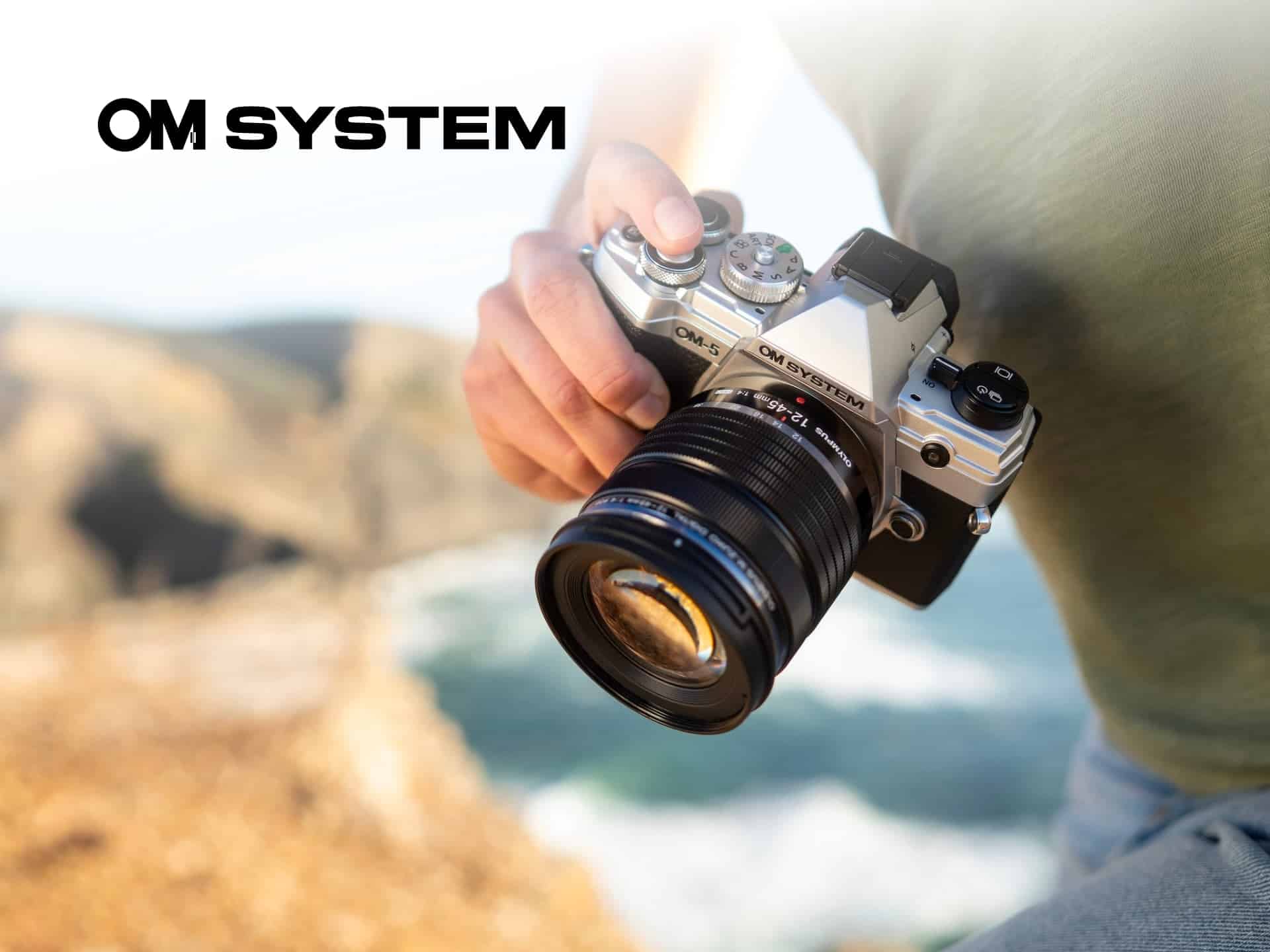 A Person Holding An Om System Camera With A Scenic Ocean View In The Background, Showcasing The Camera'S Design And Functionality For Outdoor Photography.