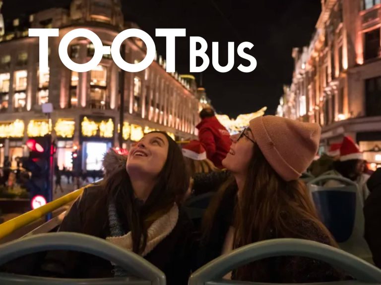 Two Friends Enjoying A Nighttime Sightseeing Tour On A Tootbus, With Festive City Lights In The Background.