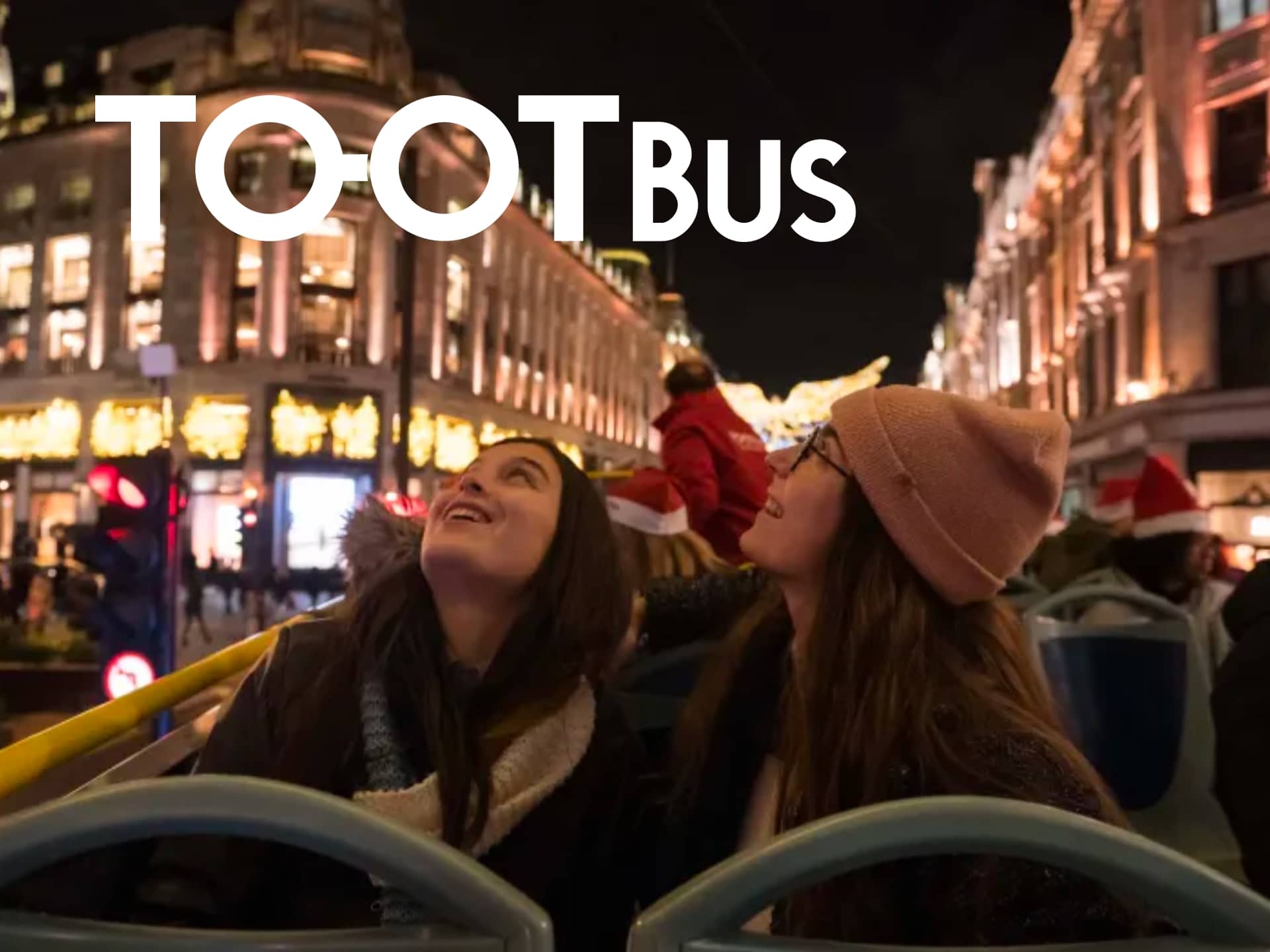 Two Friends Enjoying A Nighttime Sightseeing Tour On A Tootbus, With Festive City Lights In The Background.