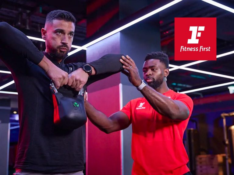 Personal Trainer Demonstrating Kettlebell Exercise With Client In Modern Gym Setting, Featuring Fitness Branding In The Background.