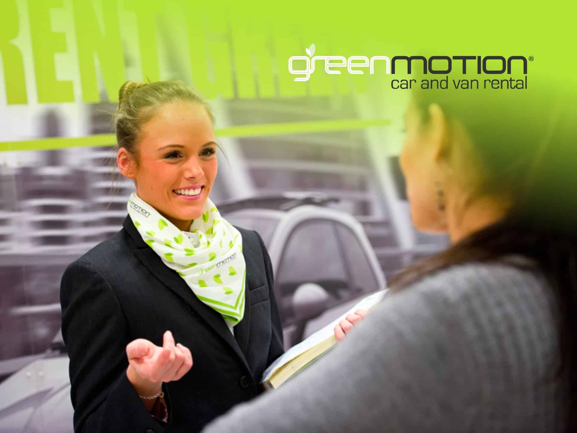 Professional Woman In A Black Suit And Green Scarf Engaging In Conversation At A Green Motion Event, Highlighting Eco-Friendly Car Rental Services.