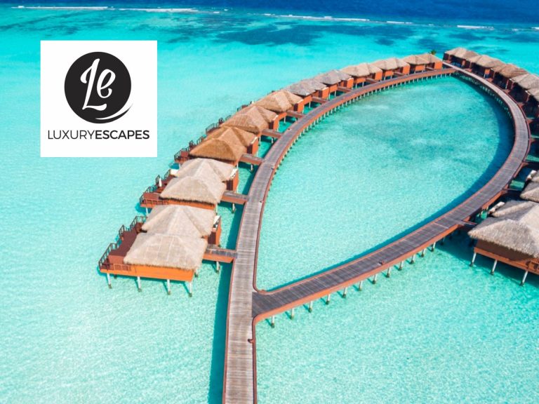 Aerial View Of Luxurious Overwater Bungalows Connected By Wooden Walkways In A Turquoise Lagoon, Featuring A Logo For A Luxury Travel Brand.